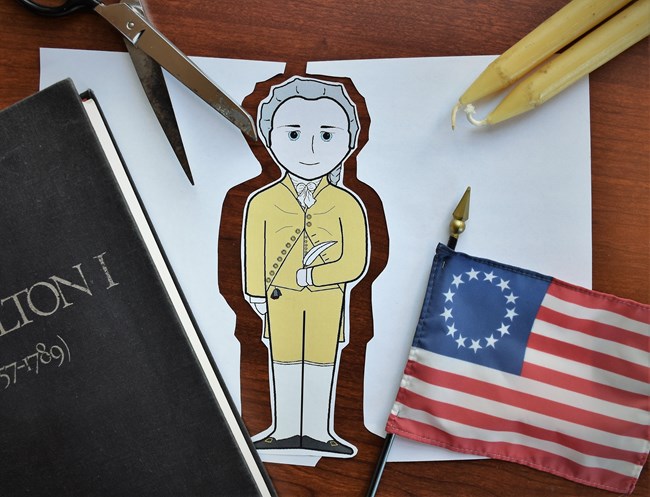 A cartoon depiction of Alexander Hamilton is surrounded by scissors, a flag, a book and candlesticks.