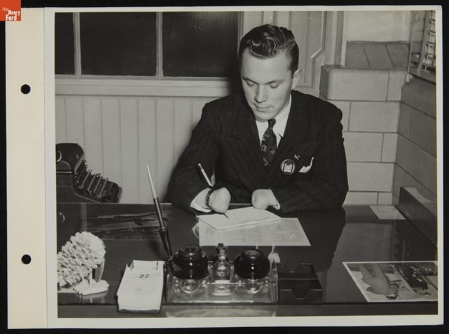 A man wearing a dark pinstriped sits at a desk. He uses a pen strapped to his forearm to write because he has stumps in place of hands.