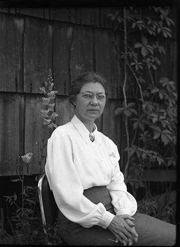 A woman wearing a dark skirt, white blouse and glasses sits in a chair facing right, her hands a clasped in her lap.