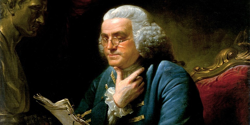 Franklin reading papers seated wearing bi-focals and blue coat.