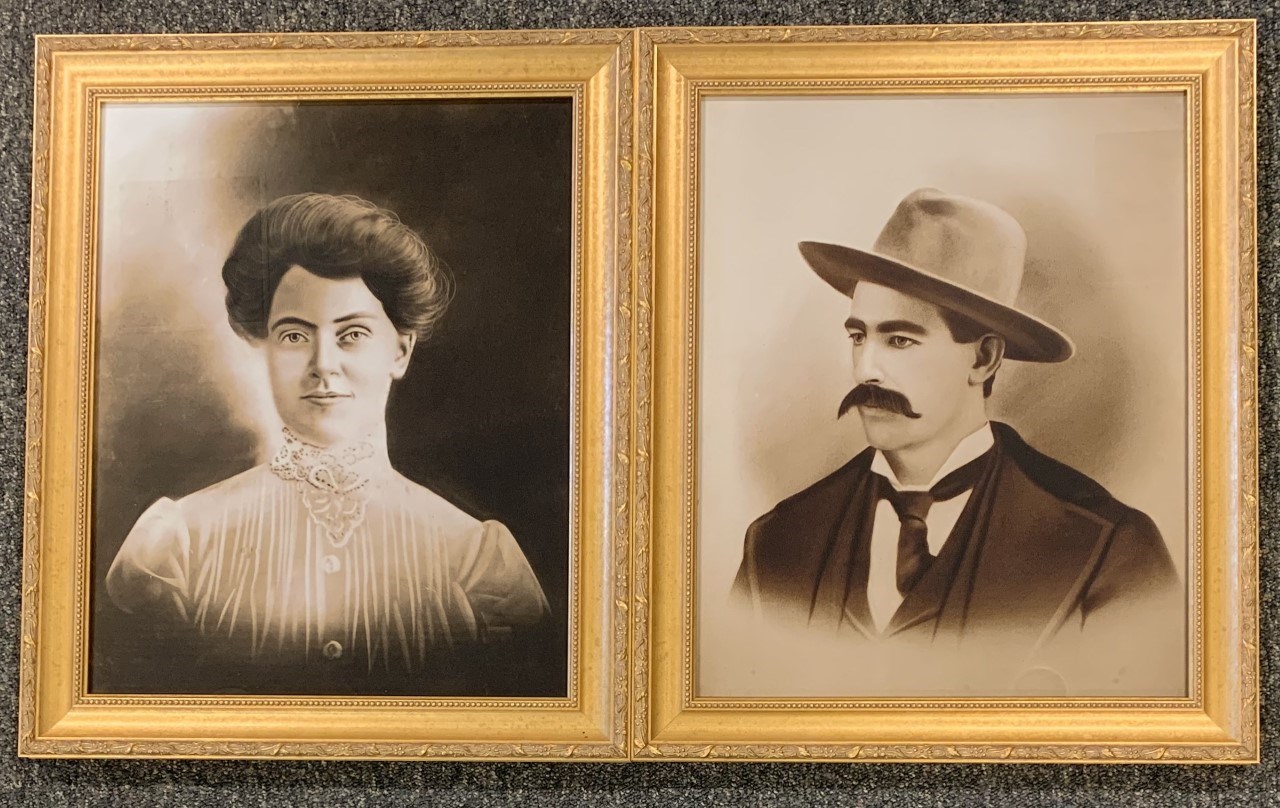 Two framed portraits of a woman on the left in a white blouse with her hair pulled up and a man on the left wearing a hat with a thick mustache.