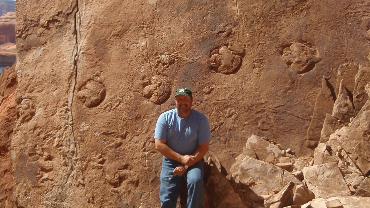 GLCA Santucci 6D269B3A-1DD8-B71B-0B6199E3E18998D6 Photo of a person sitting in front of a large boulder slab featuring fossil dinosaur tracks.