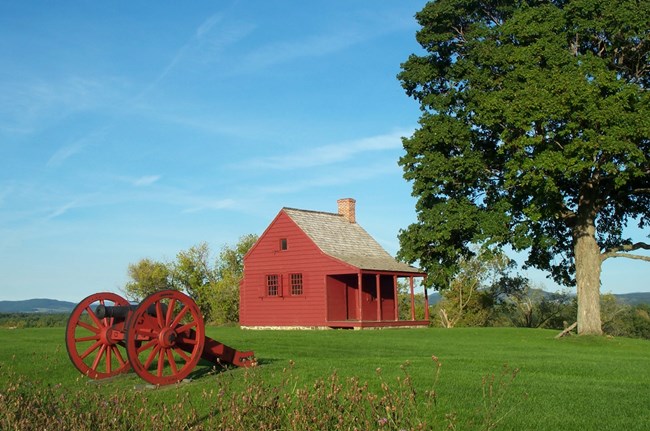 A red, two-story clapboard house sits on a verdant grass field near a line of trees and shrubs. In the left foreground, a cannon mounted on a red carriage point away from the house. Rolling caps of wooded mountains are visible in the background.