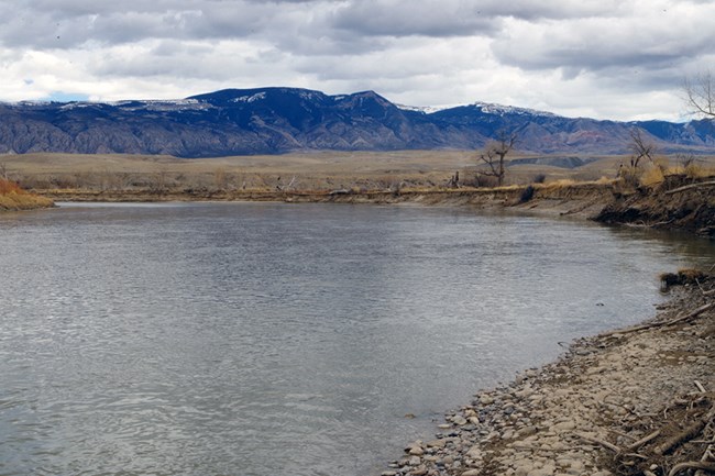 Bighorn River at Kane A bend in a river lined by rocky shores and dirt banks with snow tipped mountains in the distance.