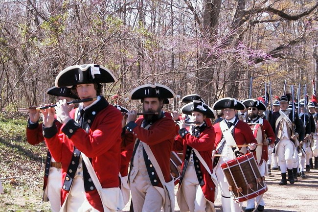 Med in historic British army uniforms with black tricorn hats, red coats, and white pants march in line playing fifes and drums