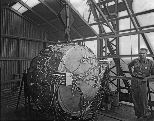 Courtesy of Los Alamos National Laboratory Black and white photo of a bomb covered in wires. A man in coveralls leans in the right of the photo. A