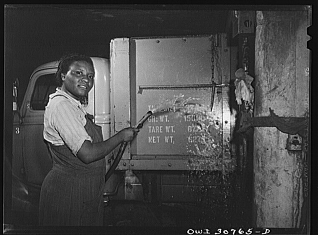 Black and white photo of African American woman in overalls, holding a hose in front of a truck