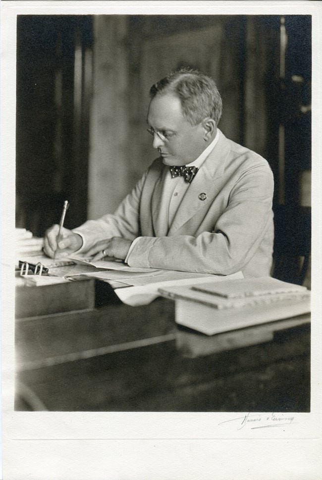 man wearing a white jacket and glasses is writing with a pencil on paper at a large desk covered in books and paper