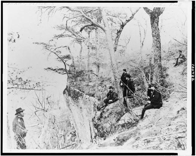 Black and white photo of a man in uniform at the bottom of a angled landscape of trees with men sitting across from him on the top of the slope