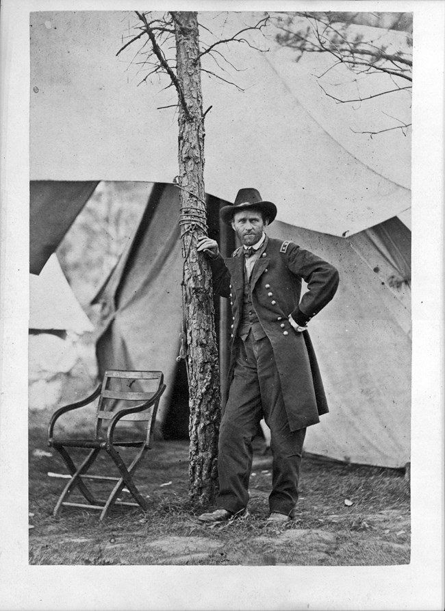 Black and white photo of a man in civil war union uniform leaning with his right shoulder on a tree with white encampments behind him