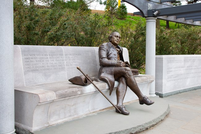 Bronze statue of George Mason seated with a book in his hand on a concrete bench with quote engraved
