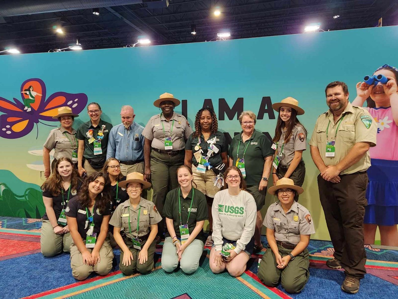 15 individuals from various federal agencies in various colored uniforms smiling in front of a sign that reads, "I Am A [Phenom]" in all caps. There is a large butterfly on the left and a girl with binoculars on the right. The floor is carpeted.
