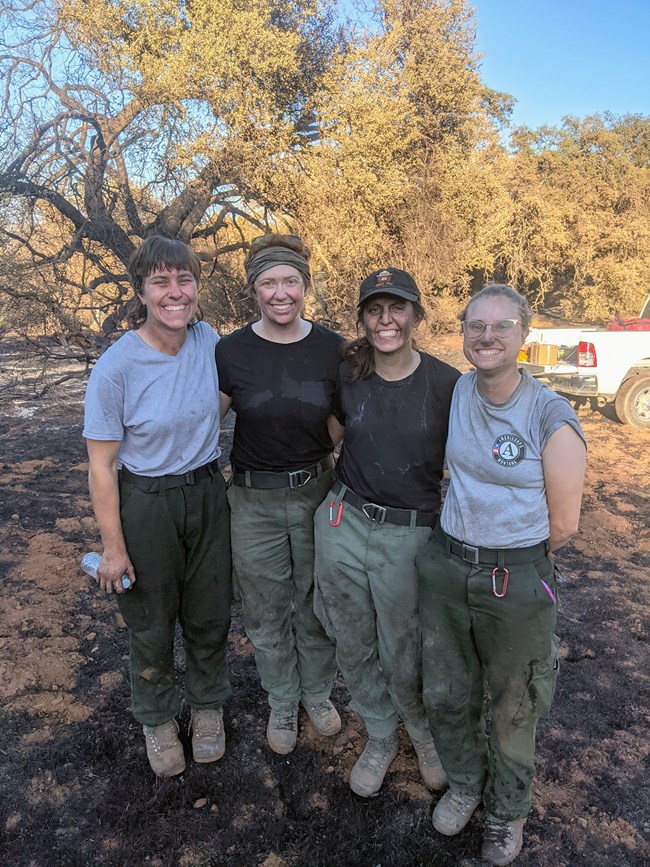 Women firefighters with dirty faces standing on charred ground.