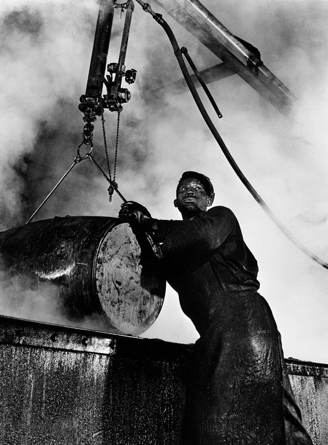 Black and white photo of a man covered in soot raising a barrel with a crane.