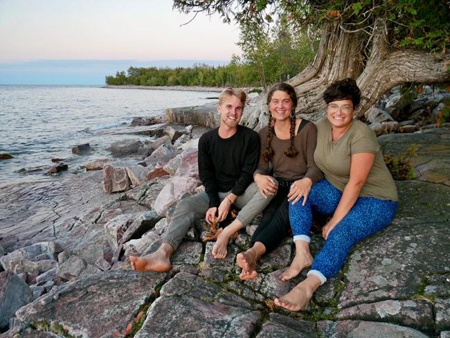 Man and two women in bare feet sit together on a rocky shoreline Man and two women in bare feet sit together on a rocky shoreline