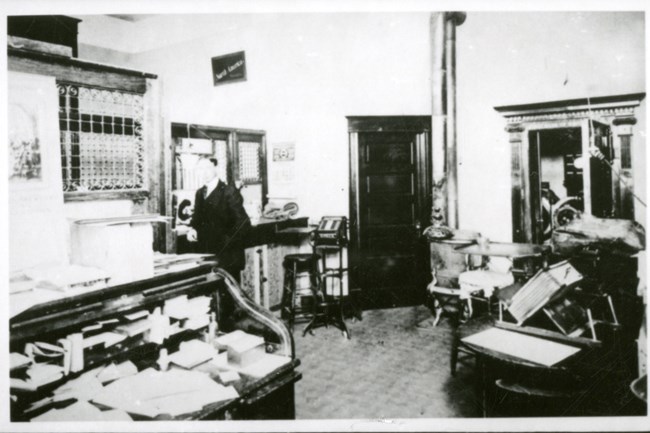 Black and white photo of the interior of a cluttered bank. A man in a suit stands at the center-left of the room.