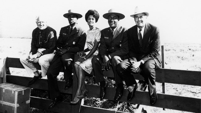 Four men and a woman sitting on a fence. The men include two park rangers in uniform and actor Henry Fonda.