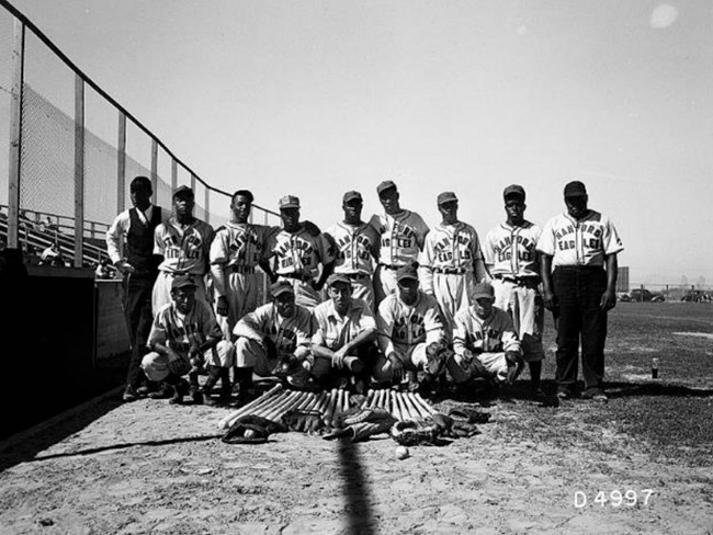 Black and white photo of African American men in baseball uniforms in two rows. Bats and gloves are laid out in front of them.