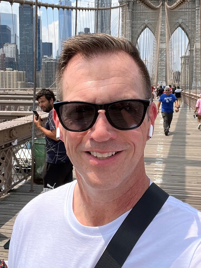 A closeup of a man in sunglasses with people walking and bridge infrastructure in the background.