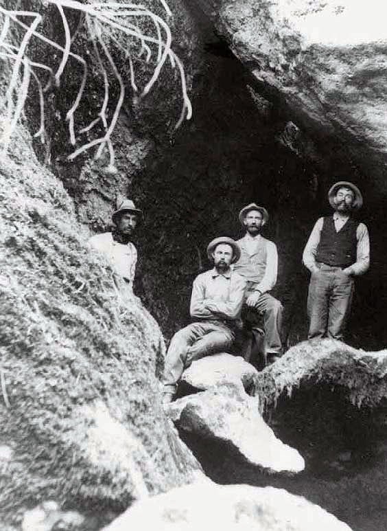 Black and white photo of four men in suit vests, jeans, and hats pose on rocks in mouth of cave.