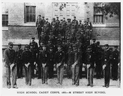 Members of DC's High School Cadet Corps, in uniform, standing in front of M Street High School in 1895. The men in the front row have their swords drawn and pointed down.