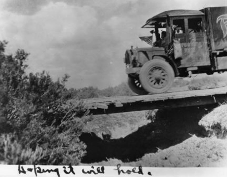 A heavy truck crosses a low ravine on a makeshift wooden bridge. Text hand-written under the photograph reads, "Hoping it will hold."