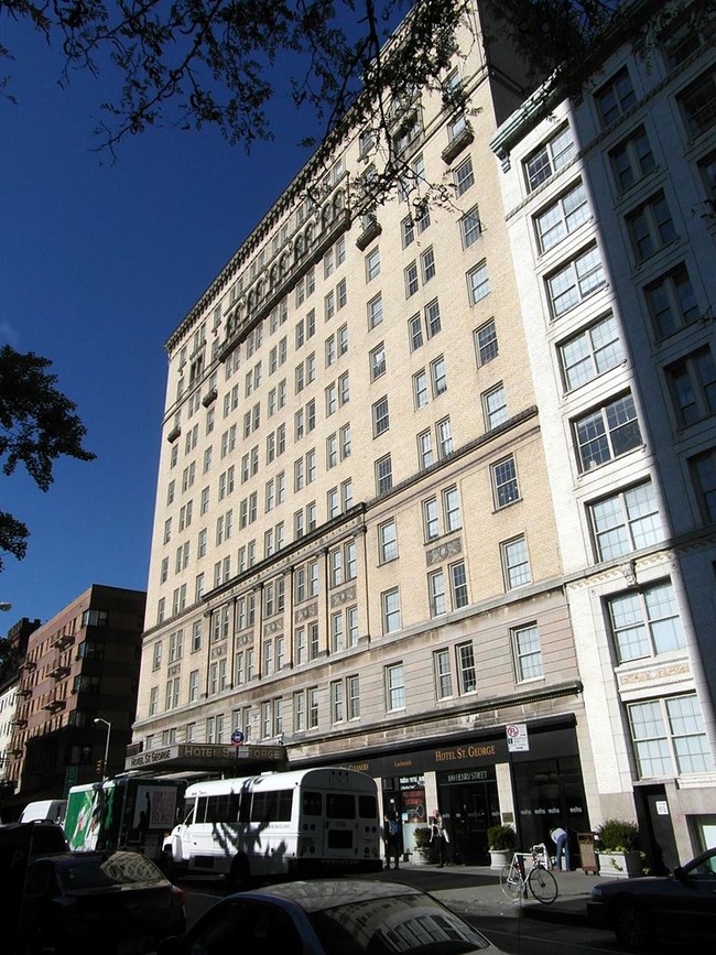 Street level view of twelve story, cream brick building with regularly spaced windows. There is a shuttle bus parked out front.
