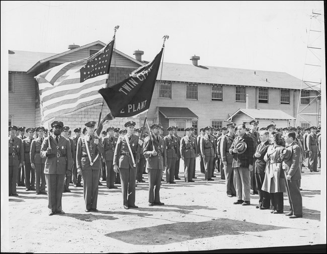 Group of soldiers in dress uniforms stand at attention in front of barracks building. In the foreground, two holding rifles flank two soldiers with American flag and TCOP flag. Three men and one woman stand to the side saluting flags.
