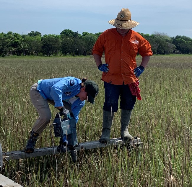One person using a drill while another watches on a platform in a salt marsh.