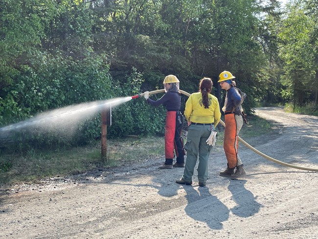 A woman in firefighter gear uses a firefighter hose, while two other women watch.