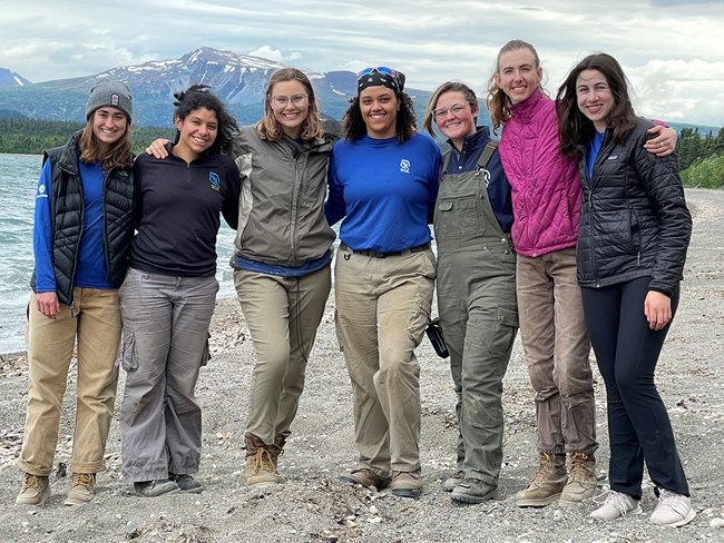 A group of seven young women smile at the camera, with a lake and mountain in the background.