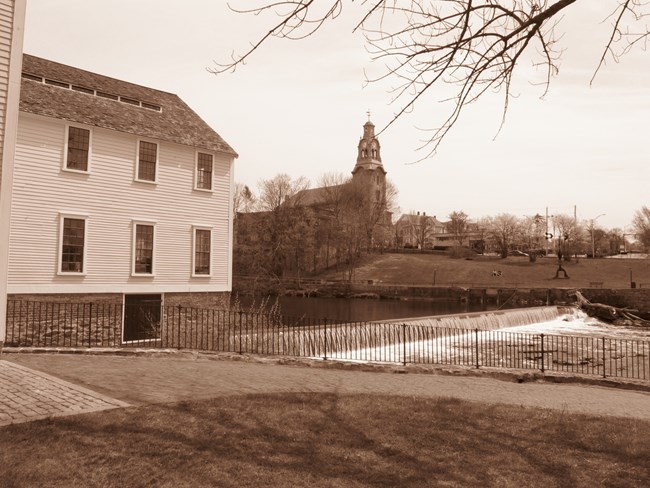 Mill building on the left of the image with dam in the center. A church stands on the hill in the background