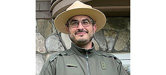 Jonathan Malzone Malzone outside of a building in a National Park Service hat and jacket, looing past the camera with a smile.