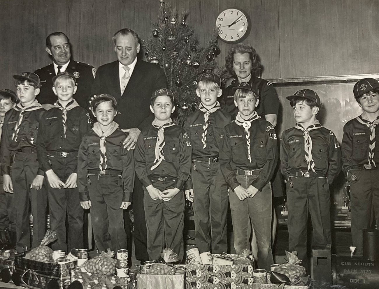 Three adults stand behind a troop of boy scouts standing in front of a train made of presents