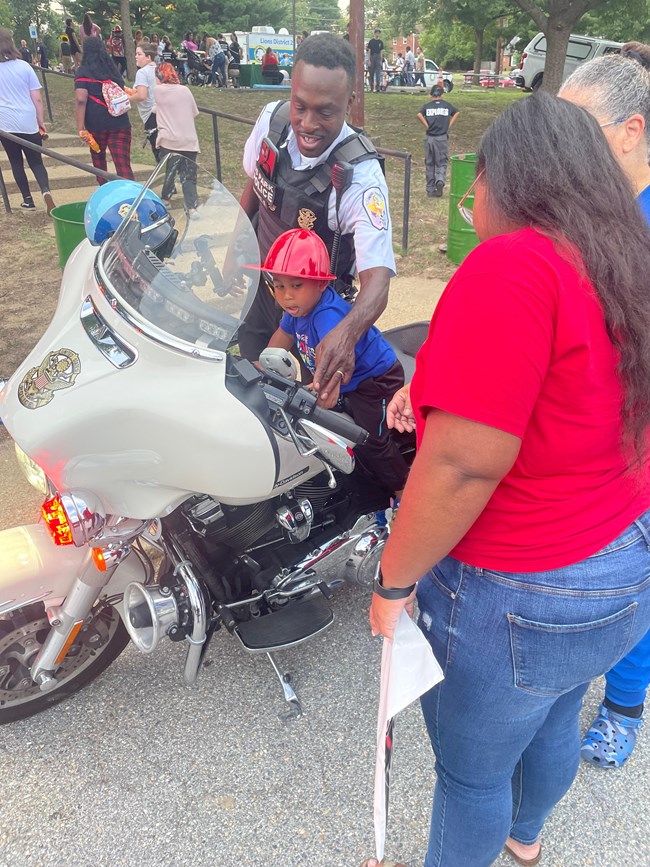 A uniformed US Park Police officer shows a boy wearing a red fireman's hat sitting on a US Park Police motorcycle how the motorcycle works. Two women watch from the side.