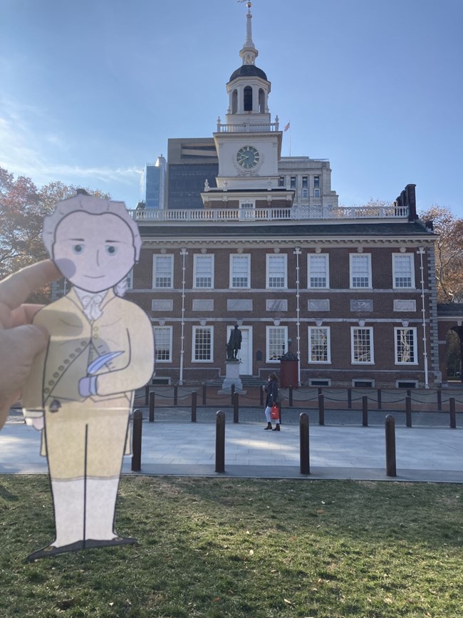 A cartoon cutout of Alexander Hamilton is held up against a background of Independence Hall.