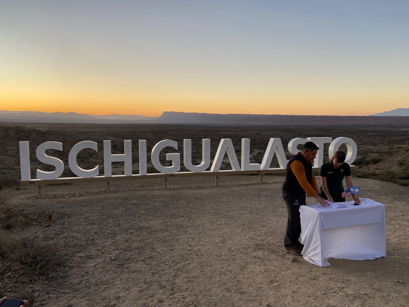 Photo two people at a small table in front of a sign for Ischigualasto.