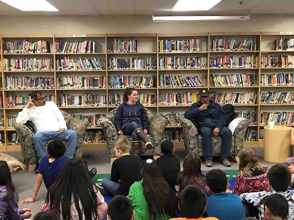 Three speakers sit in a library addressing a group seated on the floor in front of them.