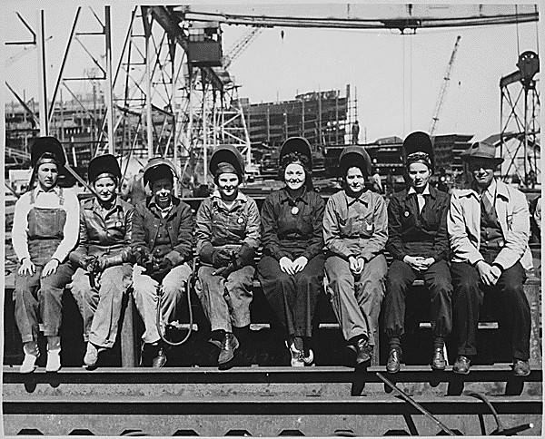 Black and white photo of eight white women in coveralls and weling helmets