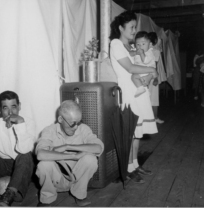 Woman holding baby stands next to radiator where two men squat. They are in a large room, divided by hanging sheets.