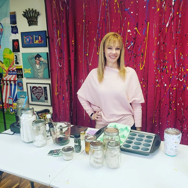 A woman with blonde hair smiles at us while standing at an art table.