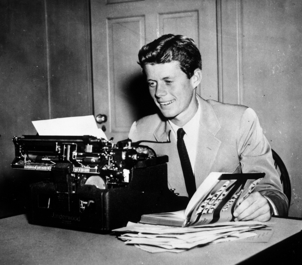 A black and white photo of John F. Kennedy in a suit, seated in front of a typewriter at a desk, smiling and holding open a book labeled ‘Why England Slept’ on the cover, which sits on a pile of papers.