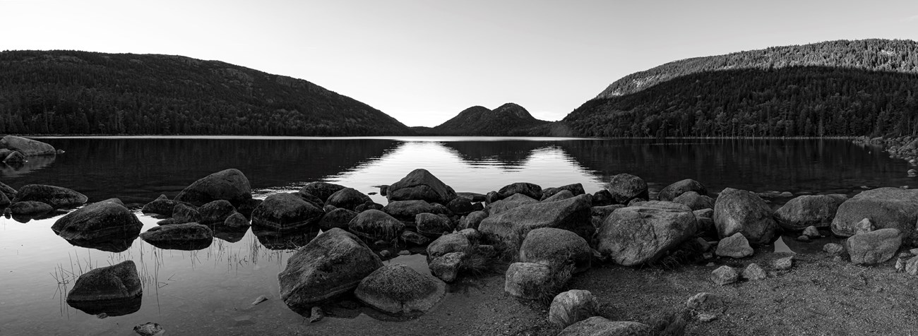 Black and white panoramic landscape photo with mountains in distance, a pond with ripple reflections, and boulders in the foreground