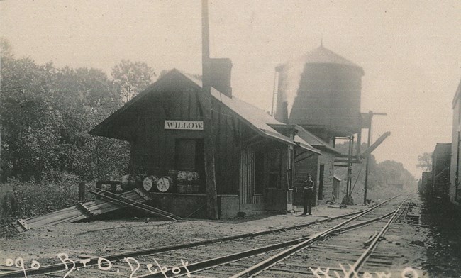 JS-13 Willow Depot looking south. Located in Independence. Modern mile post 67.5 looking at 480 bridge Black and white photo of a uniformed man standing outside the “Willow” train station. Barrels wait on the front platform. A water tower looms in the back. Text reads “090 B+O Depot, Willow, O.”