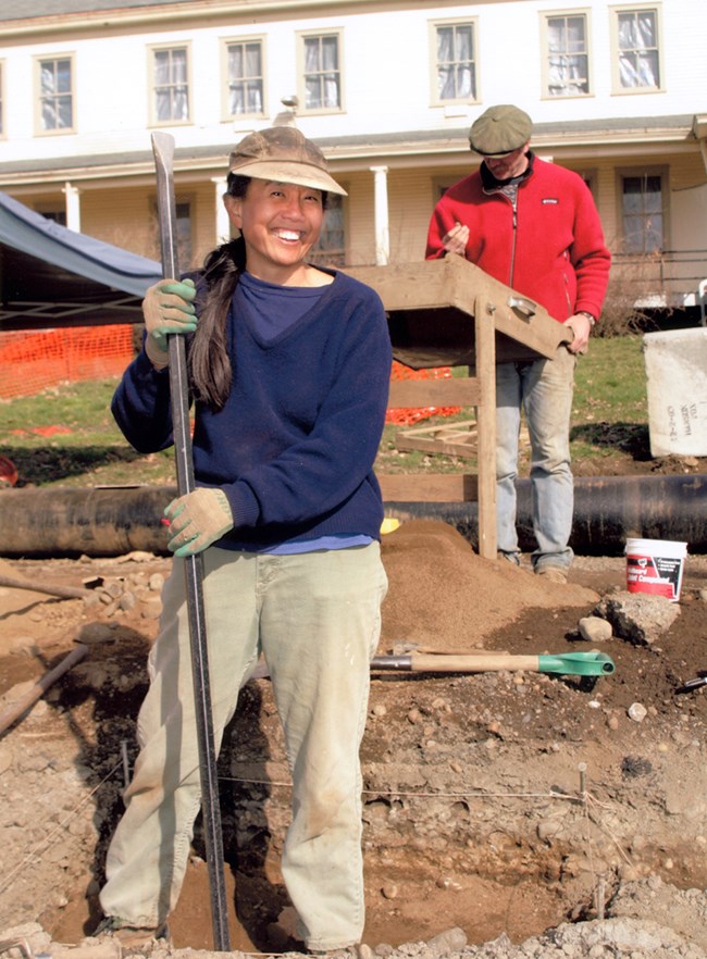 Photo of a woman holding an archaeological tool and smiling at the camera.
