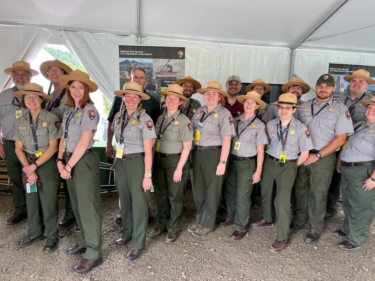 17 staff dressed in green pants and gray shirts, with gold badges, and the NPS logo. Most are wearing a flat hat with some in ball caps, and one without a hat.