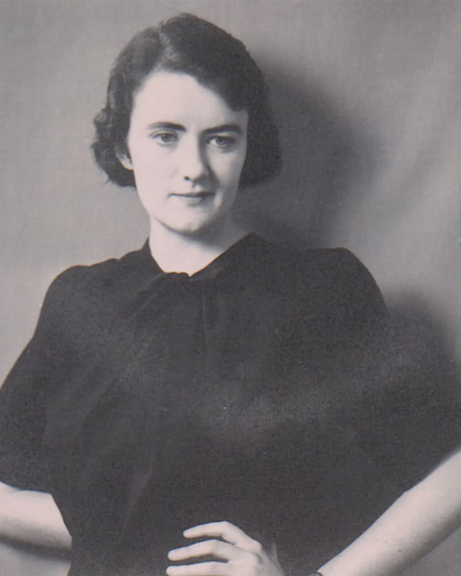 Black and white photo of a young woman in a black blouse. Her left hand is on her left hip as she flashes a small smile.