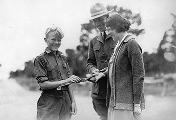 A man in a ranger uniform and smiles at his wife and son who each hold on to a stick.