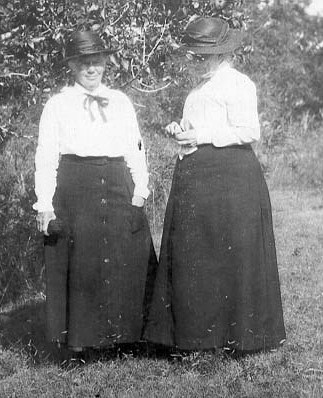 Jessie Jack and Elizabeth Owen stand in a grassy patch in front of a bush. Both wear ankle-length black skirts, white long-sleeved blouses, and black hats.