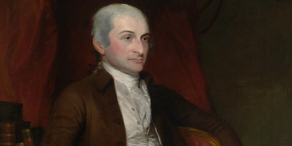 John Jay Jay in brown coat seated next to books with a red background.